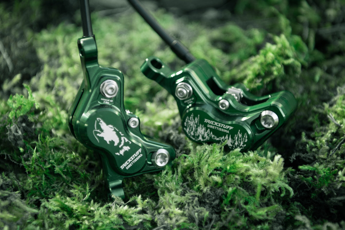 Two green Trickstuff Direttissima calipers with wolf and forest graphics resting on moss.